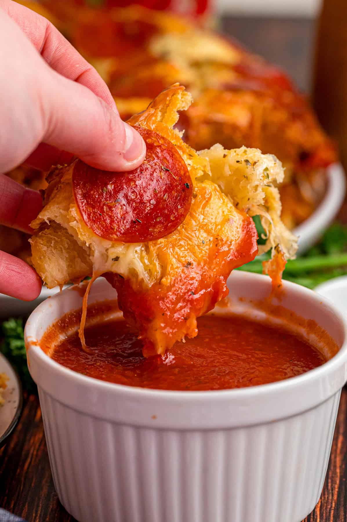 Made with biscuits, mozzarella cheese, and pepperoni, Pizza Pull Apart Bread is both easy to make and fun to eat. A hand dipping a piece of pizza pull apart bread in pizza sauce