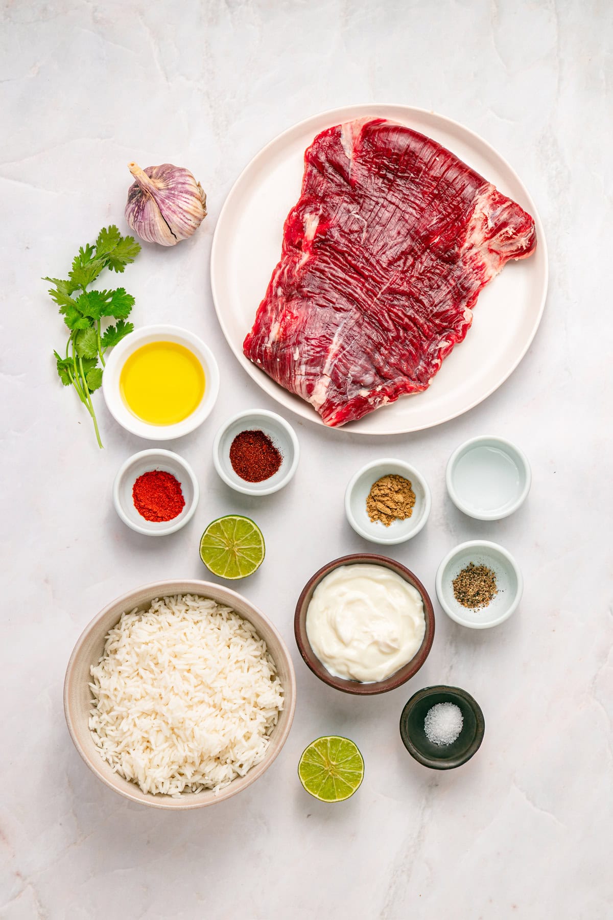 A bed of rice is topped with juicy, lightly charred flank steak, creamy yogurt lime drizzle, and veggies in this Steak Rice Bowl recipe. Overhead view of ingredients needed to make steak rice bowls