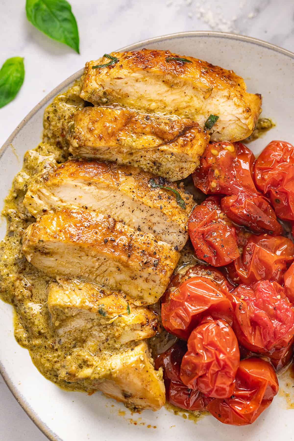 Juicy chicken breasts are cooked in a creamy pesto sauce with roasted tomatoes in this Pesto Chicken recipe. A plate of sliced pesto chicken and roasted tomatoes