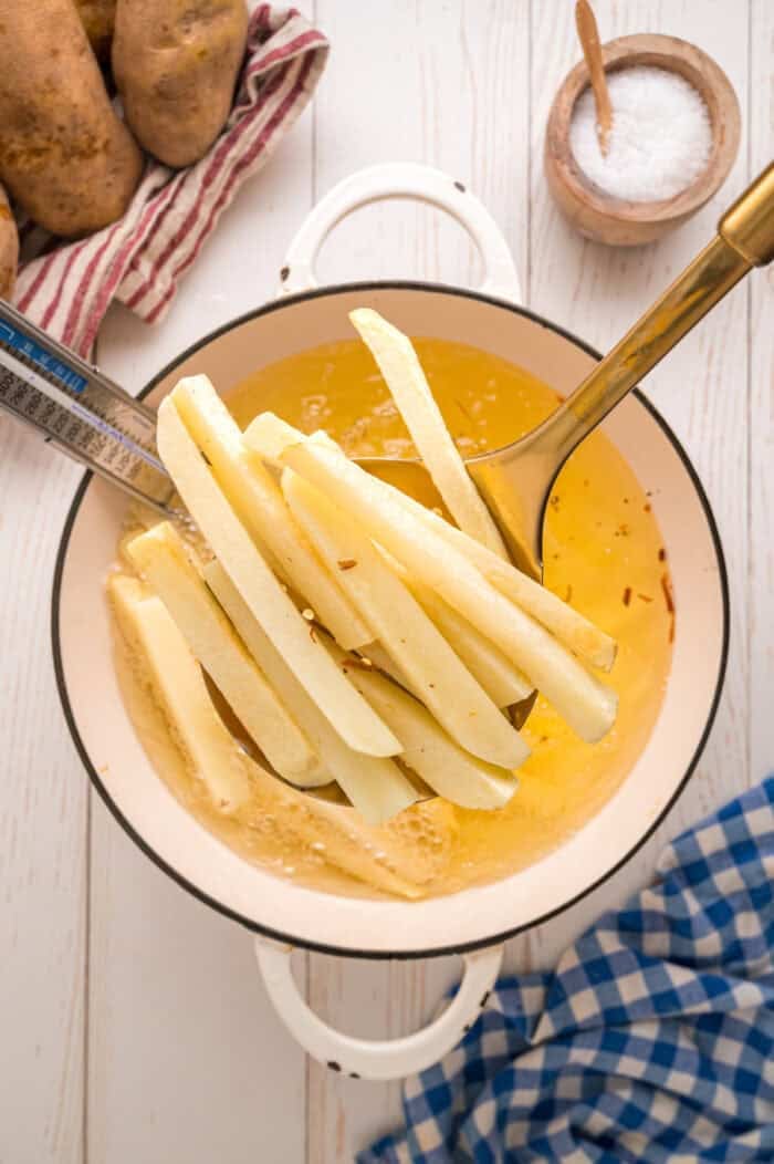 These easy homemade French Fries are made with just Russet potatoes, salt, and oil. Tender on the inside and crispy on the outside! A slotted spoon removing partially cooked fries from the oil