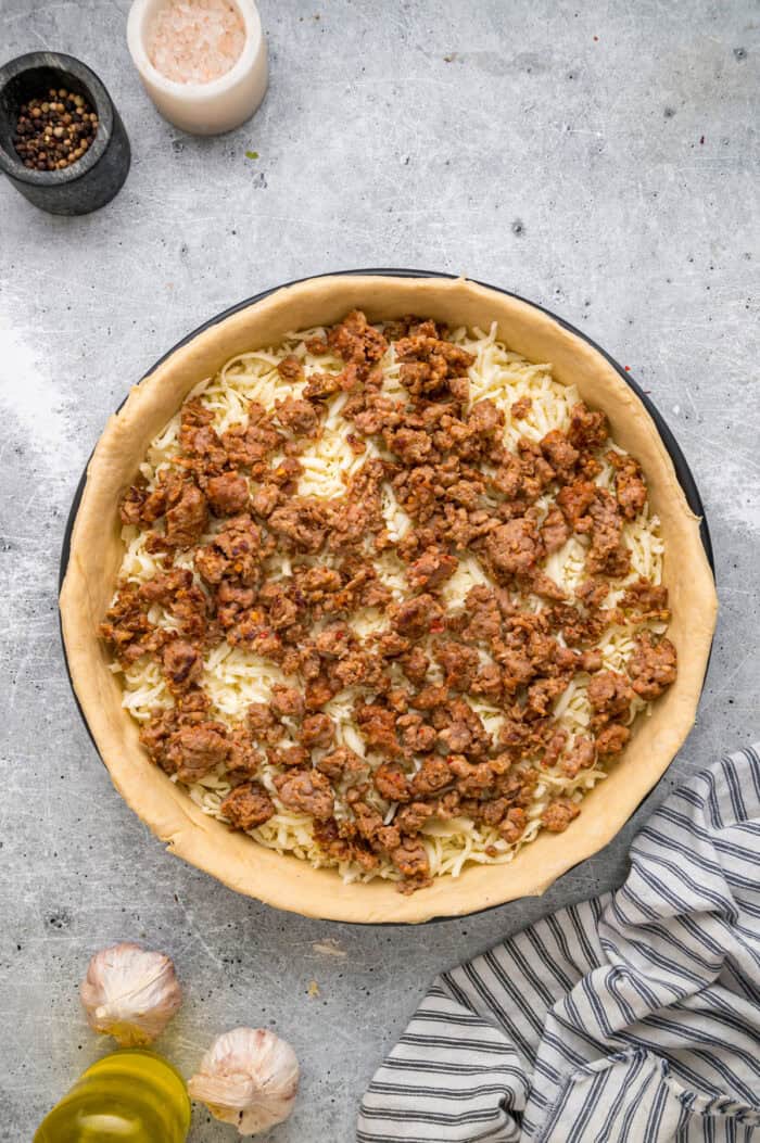 This Chicago-Style Deep Dish Pizza recipe features a buttery, flaky crust topped with mozzarella, crumbled sausage, & a savory tomato sauce. Crumbled sausage on top of mozzarella cheese in pizza dough