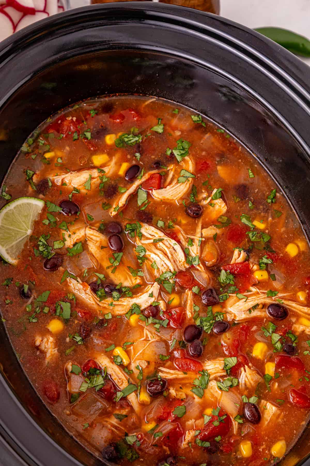 Chicken, beans, diced tomatoes and green chilies, and corn are slow-cooked in enchilada sauce broth to make Crockpot Chicken Enchilada Soup. Close up of chicken enchilada soup in the crockpot