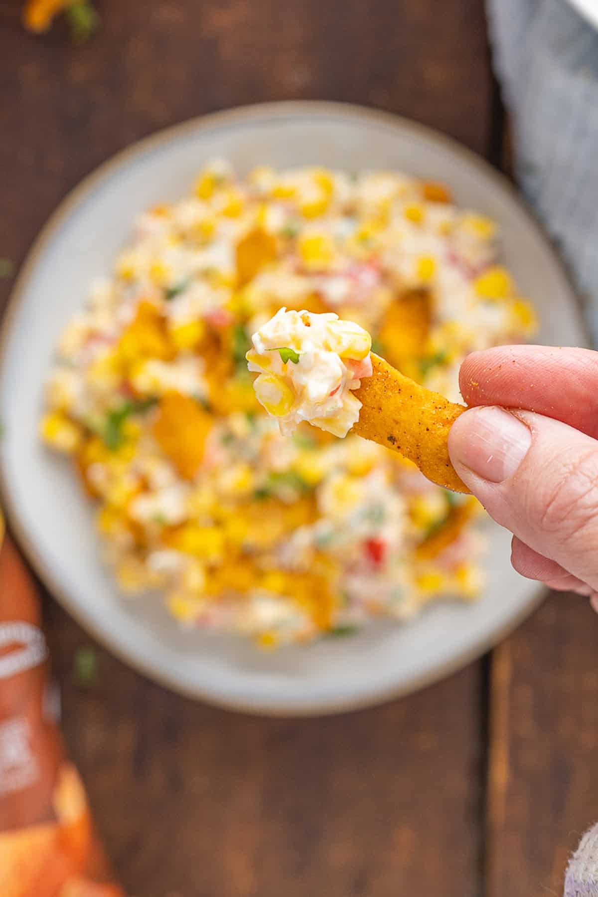 Frito Corn Salad is fun, flavorful, and the perfect combination of crunchy and creamy, with diced veggies and crushed Fritos. A hand holding a Frito dipped in corn salad