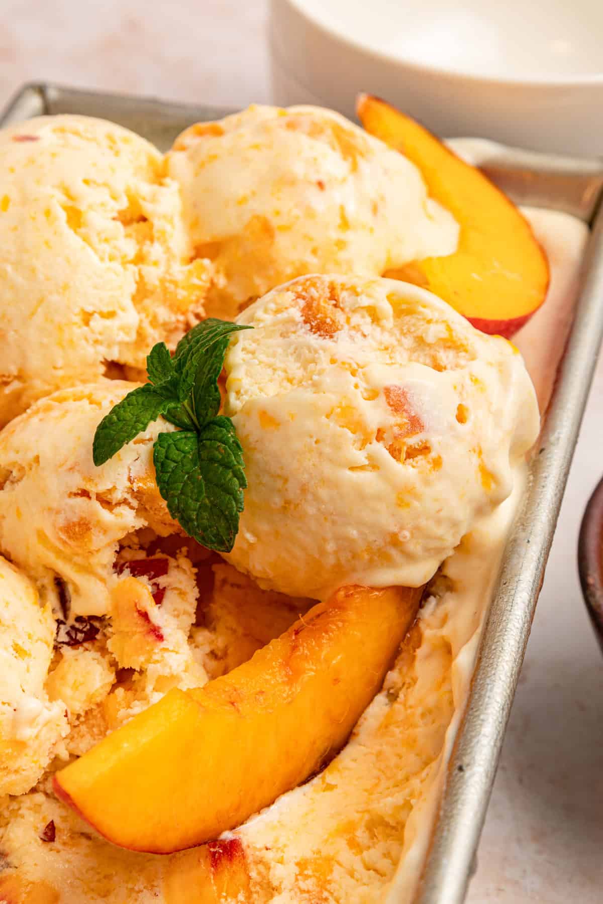 Fresh peaches are turned into a compote and swirled into a mixture of sweetened condensed milk & heavy cream in this No-Churn Peach Ice Cream. Close up of scoops of no-churn peach ice cream in the loaf pan