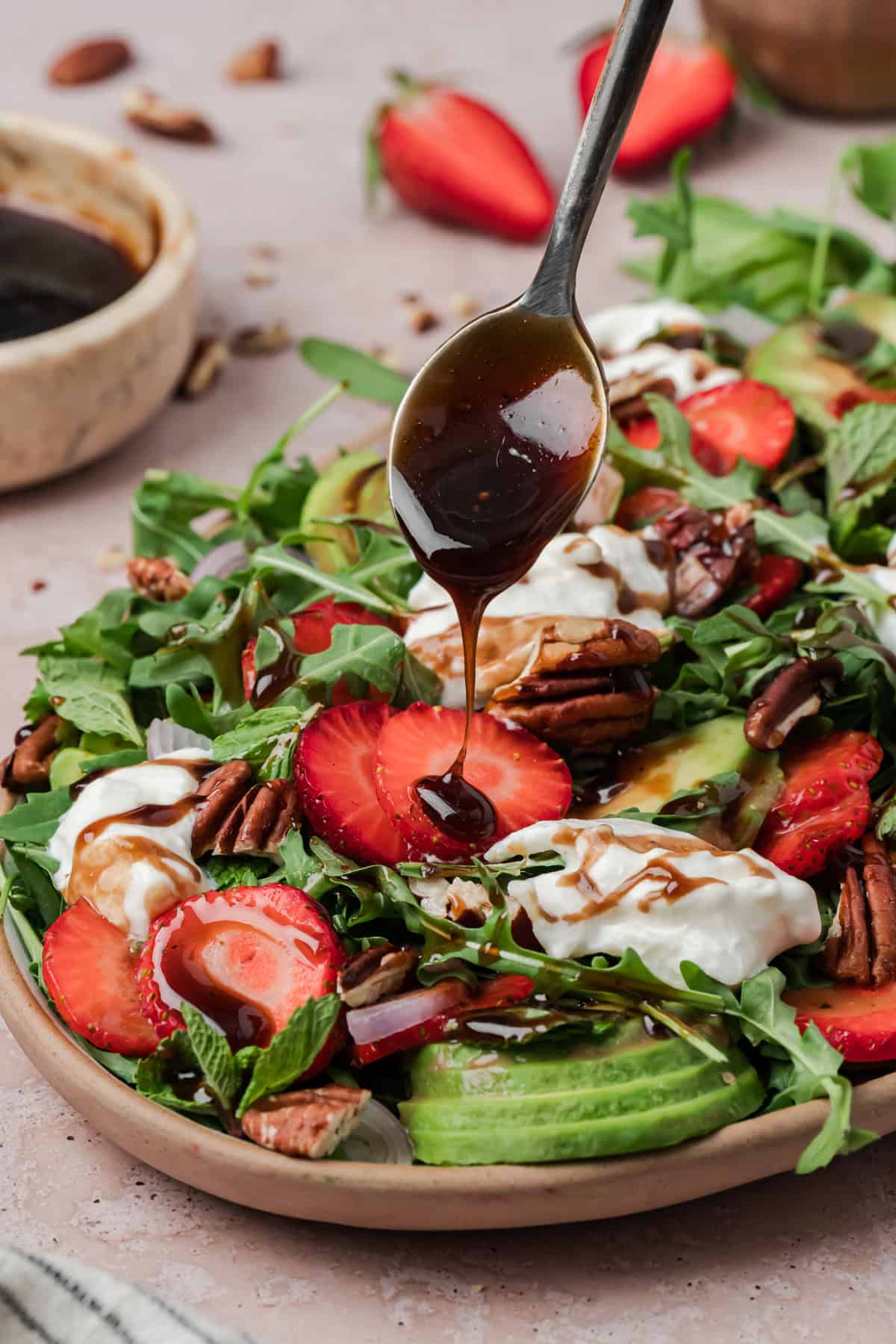 Strawberry Arugula Salad is perfect for summer, with peppery arugula, fresh strawberries, creamy burrata, crunchy pecans & avocado. A spoon drizzling balsamic vinaigrette over strawberry arugula salad