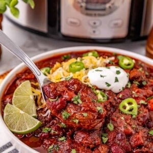 A spoon in a bowl of chile con carne in front of a crockpot