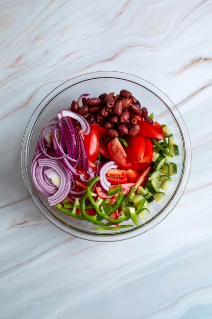 This traditional Greek Salad is made with simple ingredients & bold flavors, like fresh veggies, Kalamata olives, and feta cheese. Red onion, Kalamata olives, green peppers, and tomato in a glass bowl