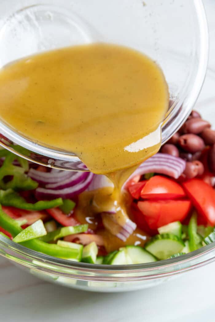 This traditional Greek Salad is made with simple ingredients & bold flavors, like fresh veggies, Kalamata olives, and feta cheese. Pouring dressing over veggies in a bowl
