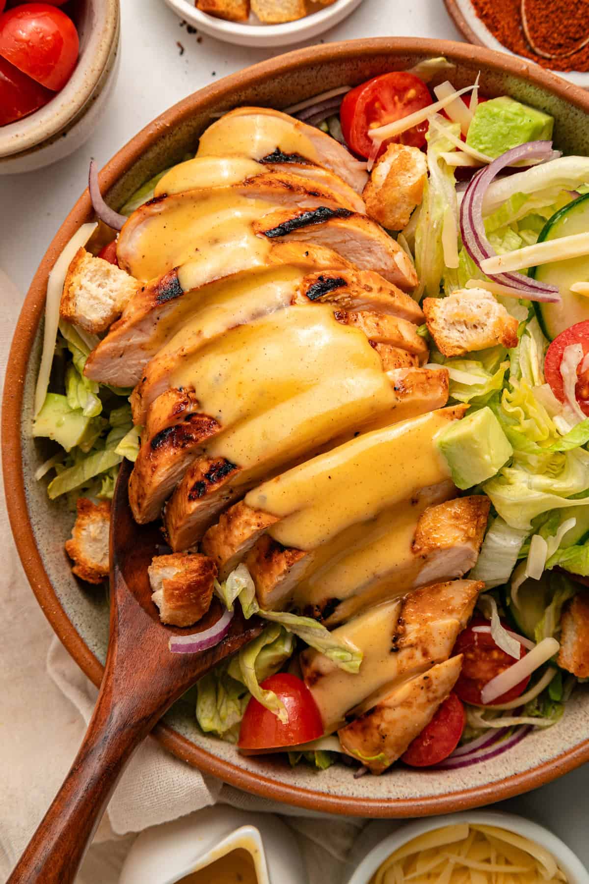 Grilled chicken sits atop a bed of lettuce with cucumber, avocado, tomato, and sharp cheddar cheese in this Honey Mustard Chicken Salad. Close up of honey mustard chicken salad in a wooden bowl with a wooden spoon
