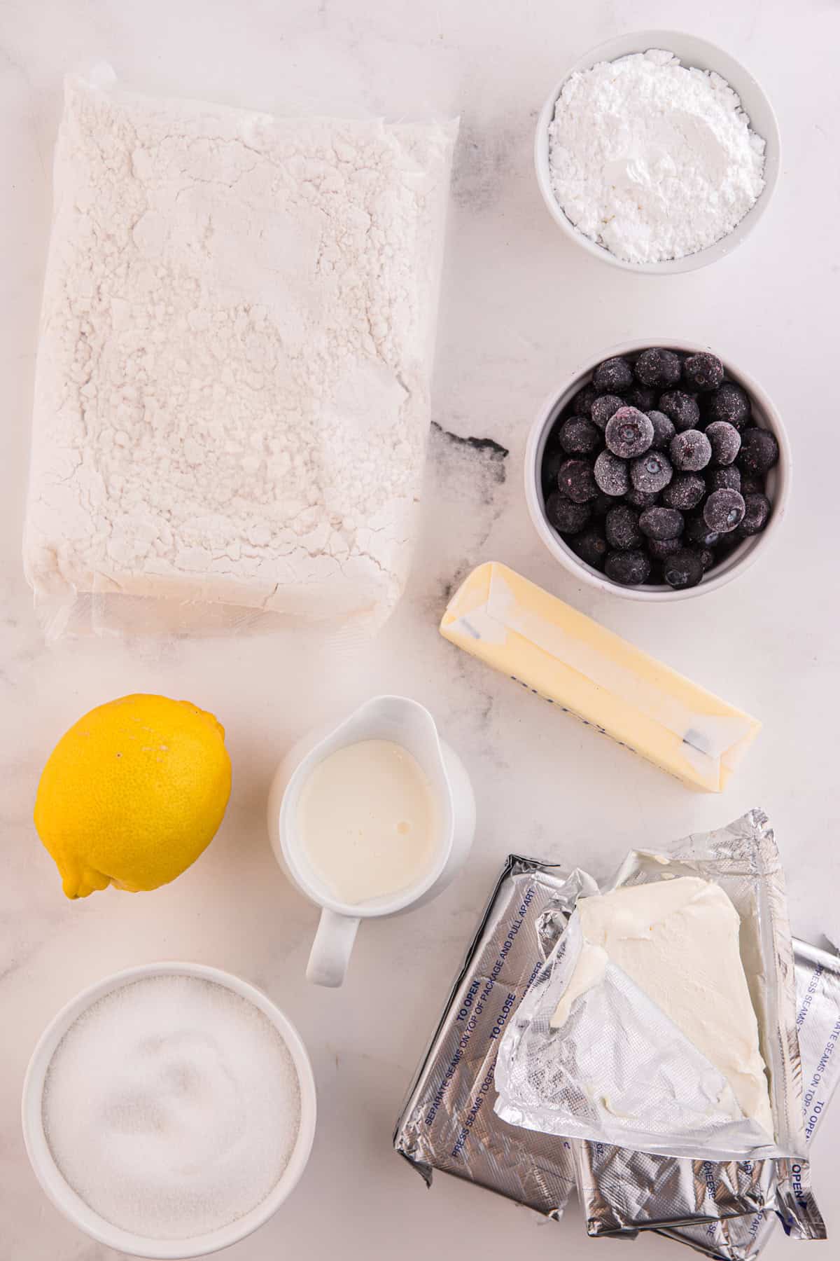 Moist lemon cakes, tangy blueberry cheesecake filling, & tart lemon frosting make up the layers of this Lemon Blueberry Cheesecake Cake. Overhead view of ingredients needed to make lemon blueberry cheesecake cake