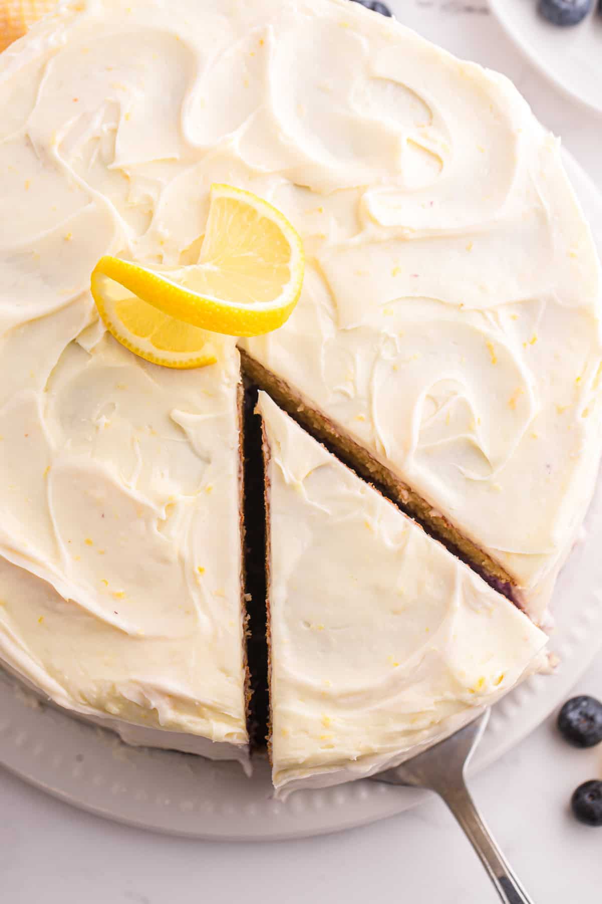Moist lemon cakes, tangy blueberry cheesecake filling, & tart lemon frosting make up the layers of this Lemon Blueberry Cheesecake Cake. Overhead view of lemon blueberry cheesecake cake