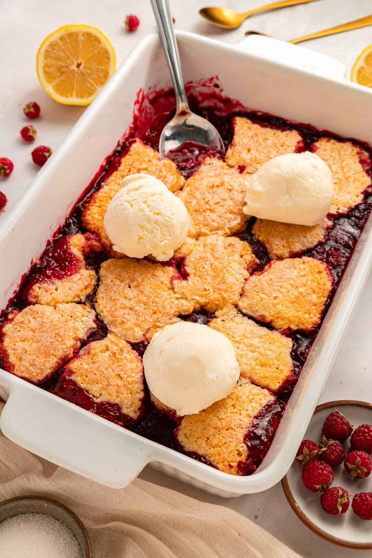 This rustic Raspberry Cobbler recipe is made with a jammy fruit filling and a golden, biscuit-style topping. Quick & easy! Overhead view of a baking dish of raspberry cobbler with scoops of vanilla ice cream
