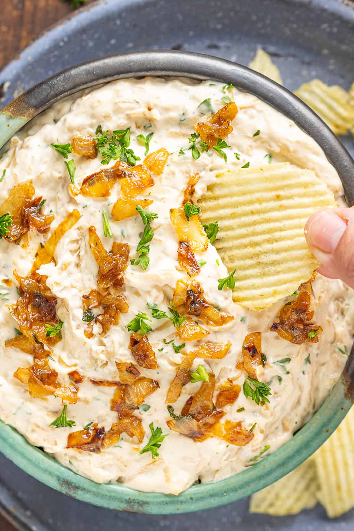 Sweet, savory caramelized onions are combined with tangy sour cream and cream cheese, plus a few seasonings, to make Caramelized Onion Dip. Overhead view of a hand dipping a chip into caramelized onion dip