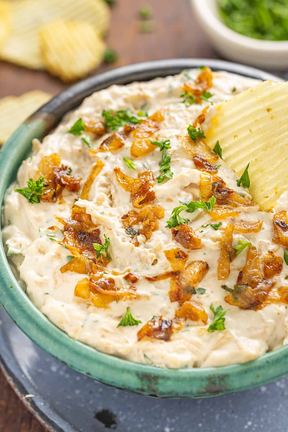 Sweet, savory caramelized onions are combined with tangy sour cream and cream cheese, plus a few seasonings, to make Caramelized Onion Dip. Close up of a bowl of caramelized onion dip with a chip