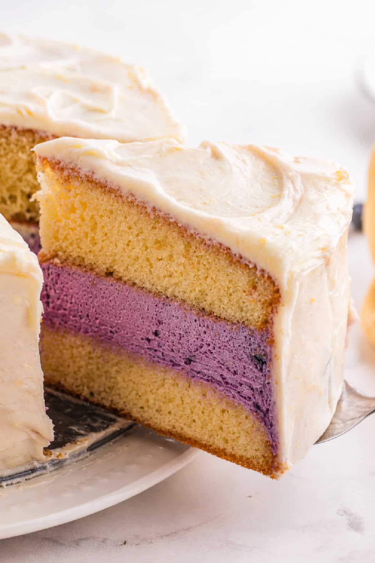 Moist lemon cakes, tangy blueberry cheesecake filling, & tart lemon frosting make up the layers of this Lemon Blueberry Cheesecake Cake. a slice of lemon blueberry cheesecake cake being lifted with a spatula