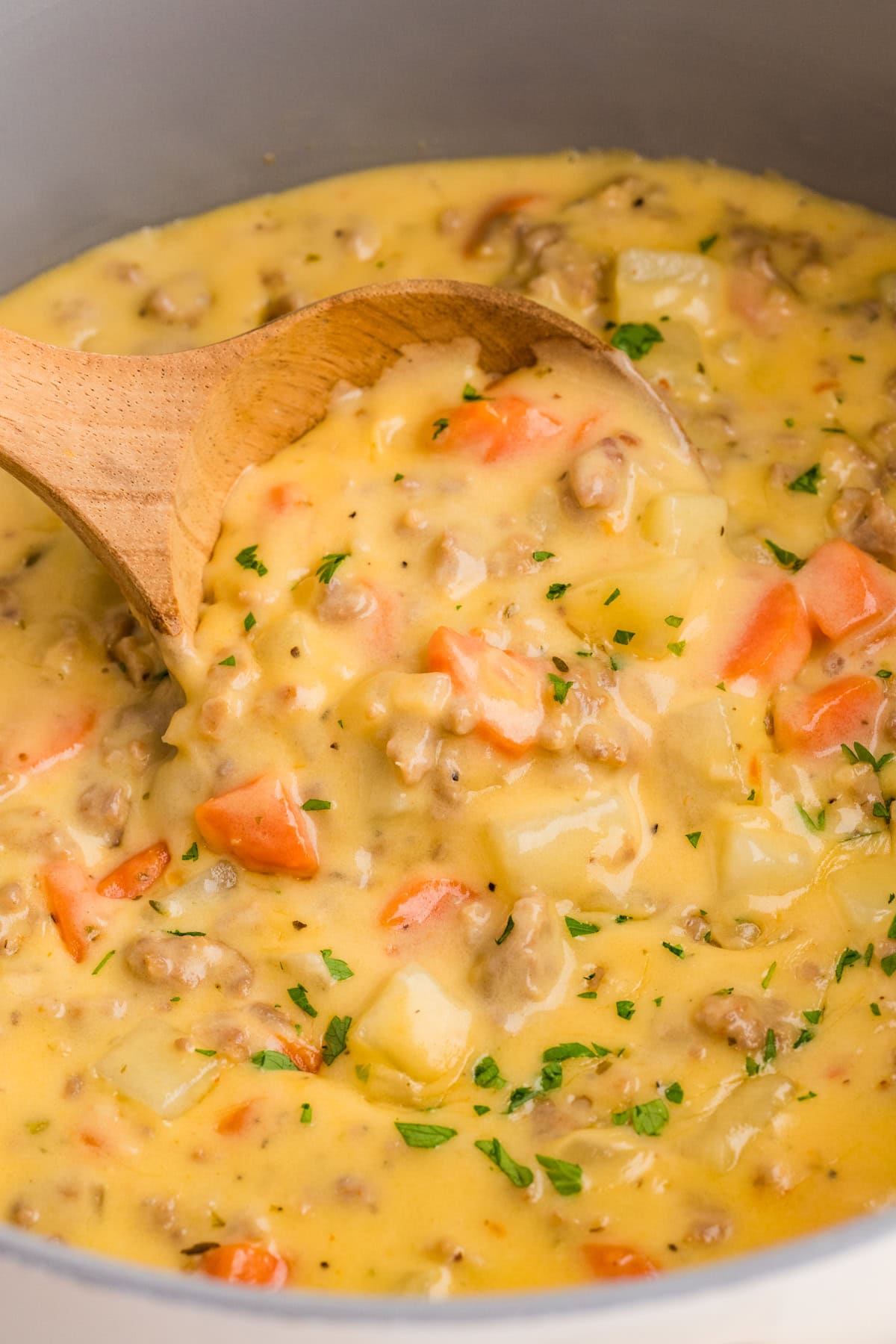 This Sausage Potato Soup is rich, creamy, and loaded with savory Italian sausage, tender potatoes, sweet carrots, and sharp cheddar cheese. A wooden spoon stirring sausage and potato soup
